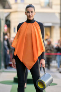 Model on a street-style runway wearing an orange draped cape over a black bodysuit, holding a gray cylindrical bag with a yellow interior.