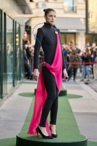 Model walking a circular green runway in a black bodysuit with a hot pink draped sash and blue accent cord, onlookers in the background.