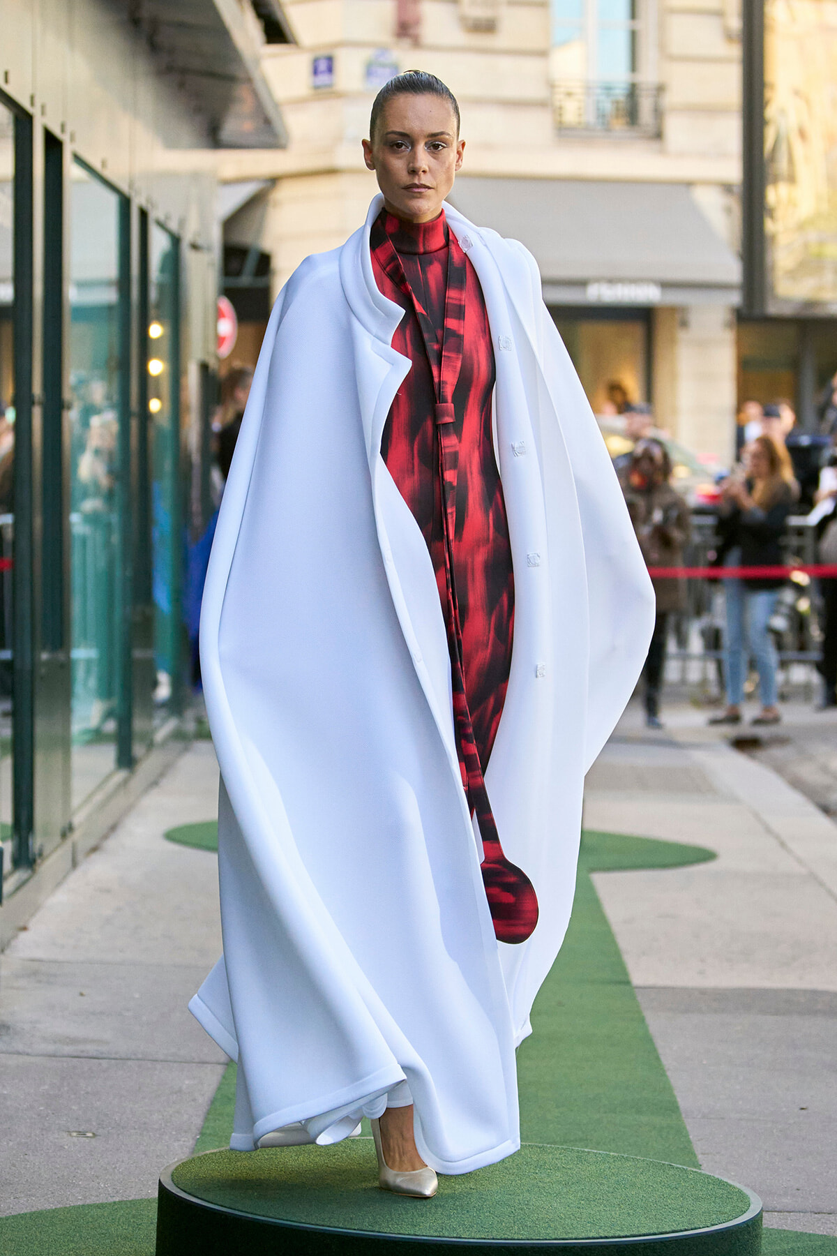 Model walks a street-style runway wearing a voluminous white cape over a red plaid dress, standing on a green circular platform.