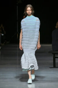 Model walking a runway in a pale blue, tiered knit dress with horizontal ruffles and a mermaid hem, paired with matching light-blue shoes.