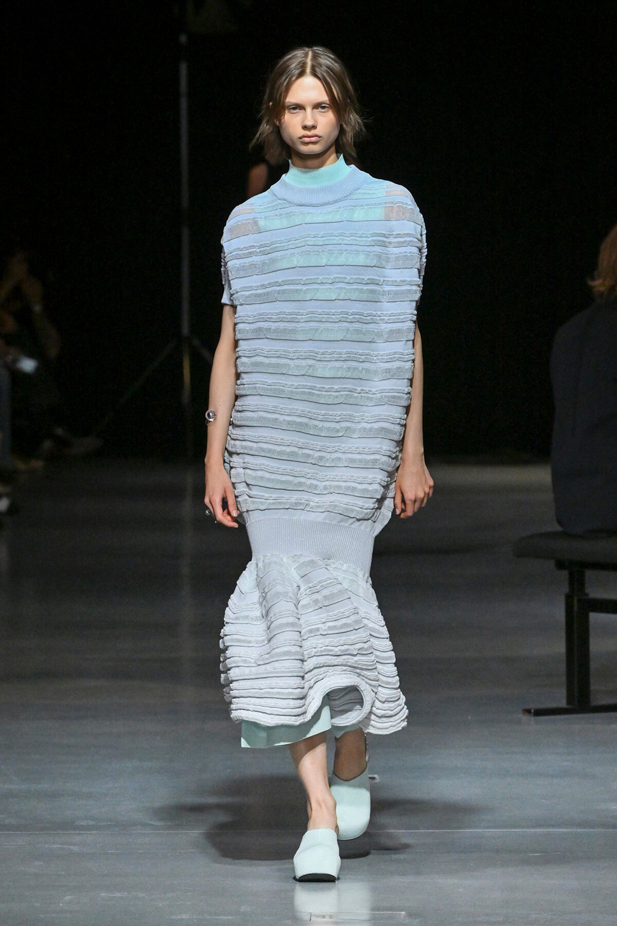 Model walking a runway in a pale blue, tiered knit dress with horizontal ruffles and a mermaid hem, paired with matching light-blue shoes.