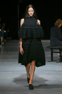 Runway model walking in a black, multi-layered dress with cold-shoulder sleeves and a high-neck top.