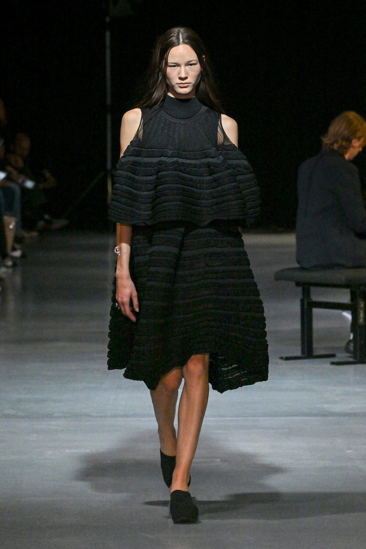 Runway model walking in a black, multi-layered dress with cold-shoulder sleeves and a high-neck top.