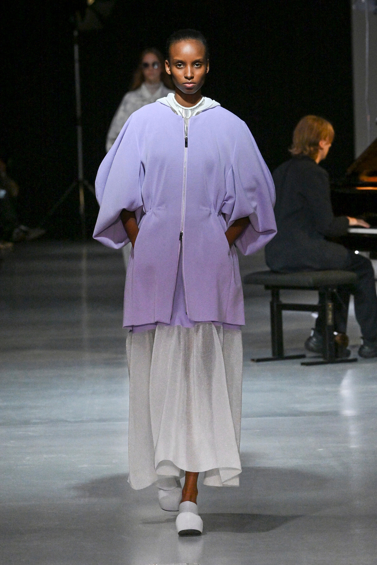 Runway model in a lavender oversized zip-front cape over a white shimmering ankle-length skirt, with white shoes; pianist in background.