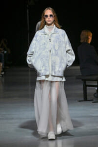 Model on a runway wearing an oversized pale blue tie-dye jacket over a sheer, floor-length white skirt, with round sunglasses.