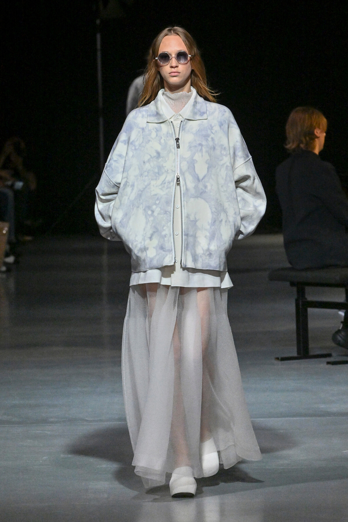 Model on a runway wearing an oversized pale blue tie-dye jacket over a sheer, floor-length white skirt, with round sunglasses.