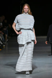 Model on a runway wearing a pale blue-gray, tiered knit dress with horizontal ruffle stripes and a high-neck top, full-length view.
