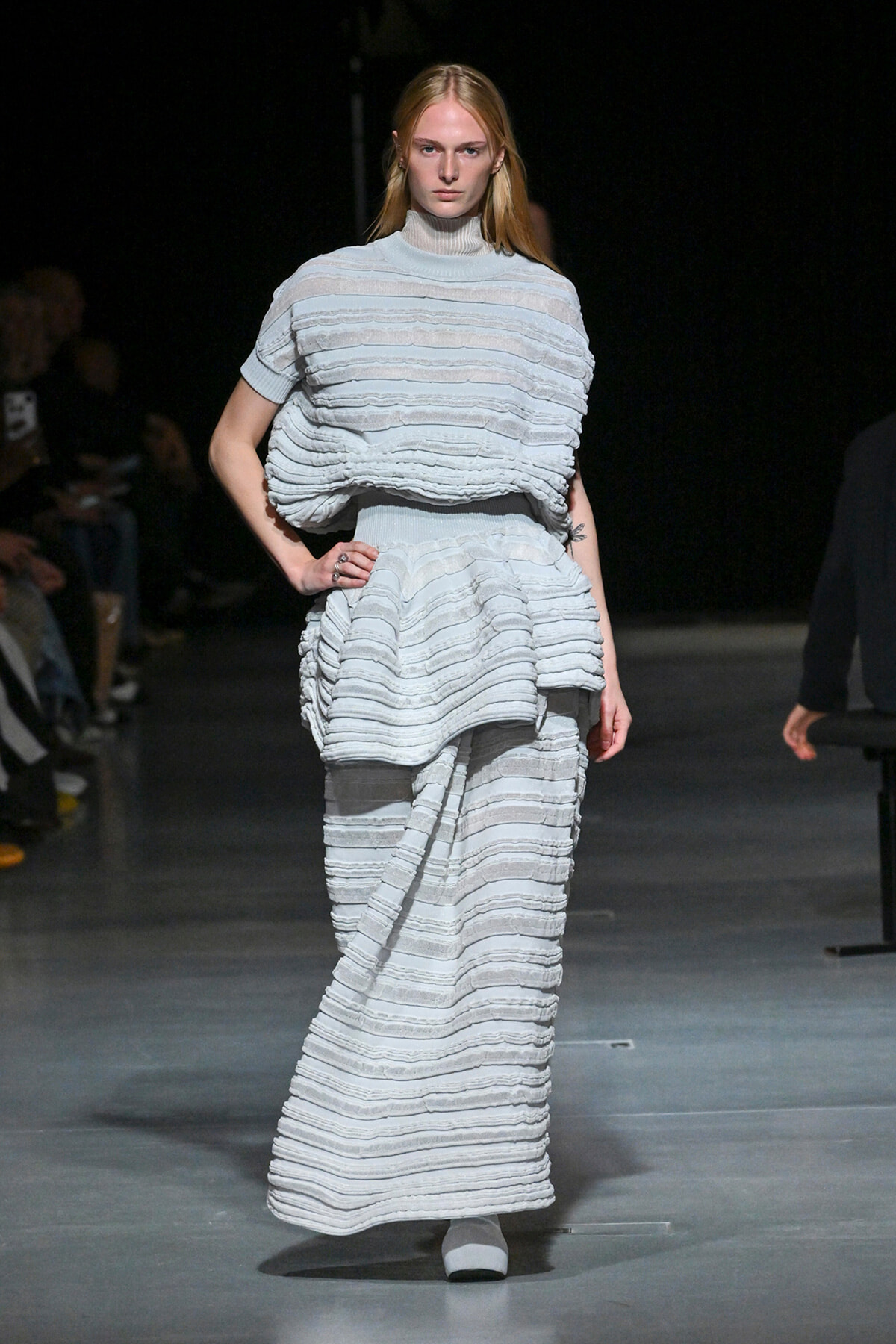 Model on a runway wearing a pale blue-gray, tiered knit dress with horizontal ruffle stripes and a high-neck top, full-length view.