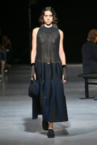Runway model wearing a sheer black sleeveless blouse with a high neckline and a navy pleated midi skirt, carrying a small dark bag with black shoes.