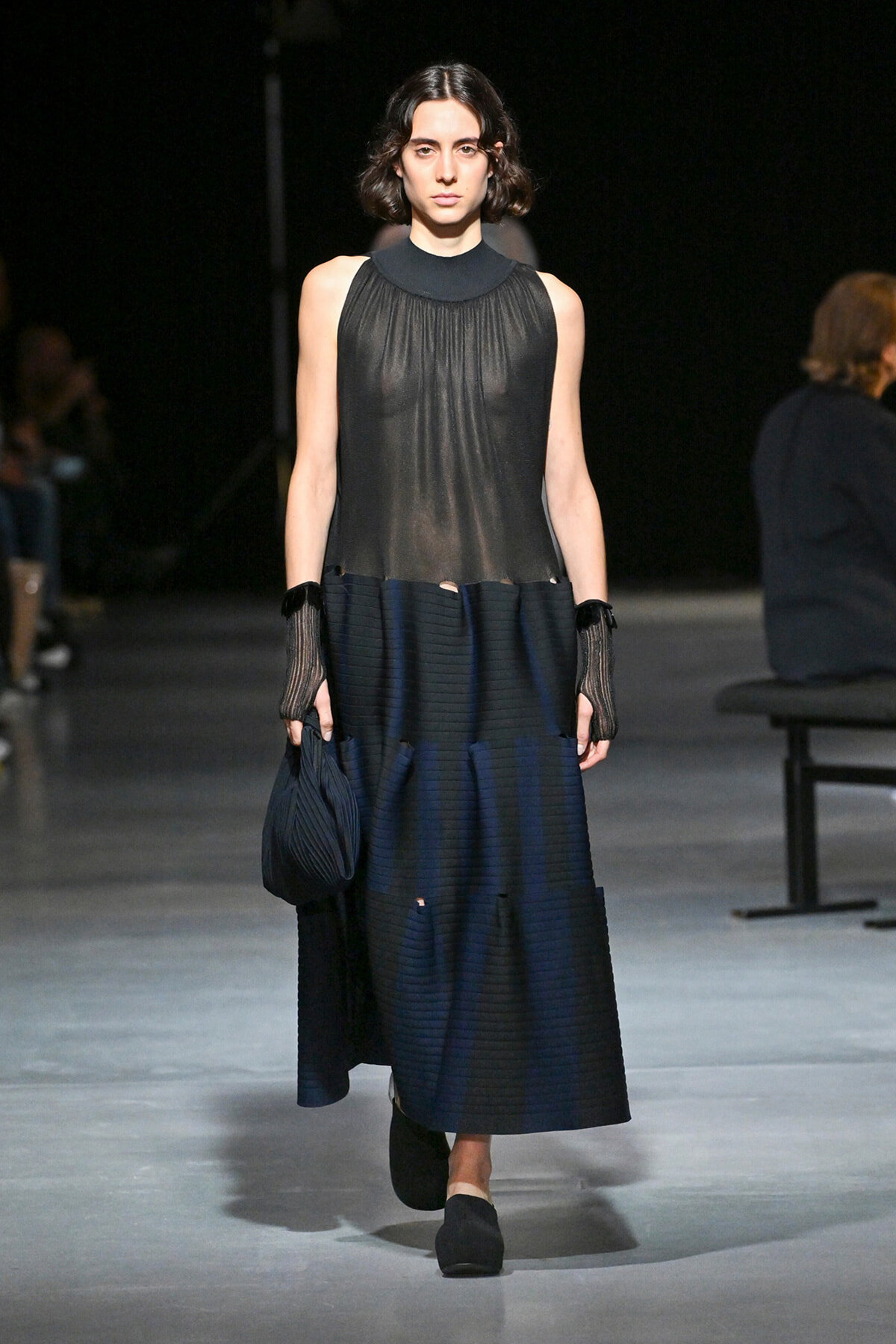 Runway model wearing a sheer black sleeveless blouse with a high neckline and a navy pleated midi skirt, carrying a small dark bag with black shoes.
