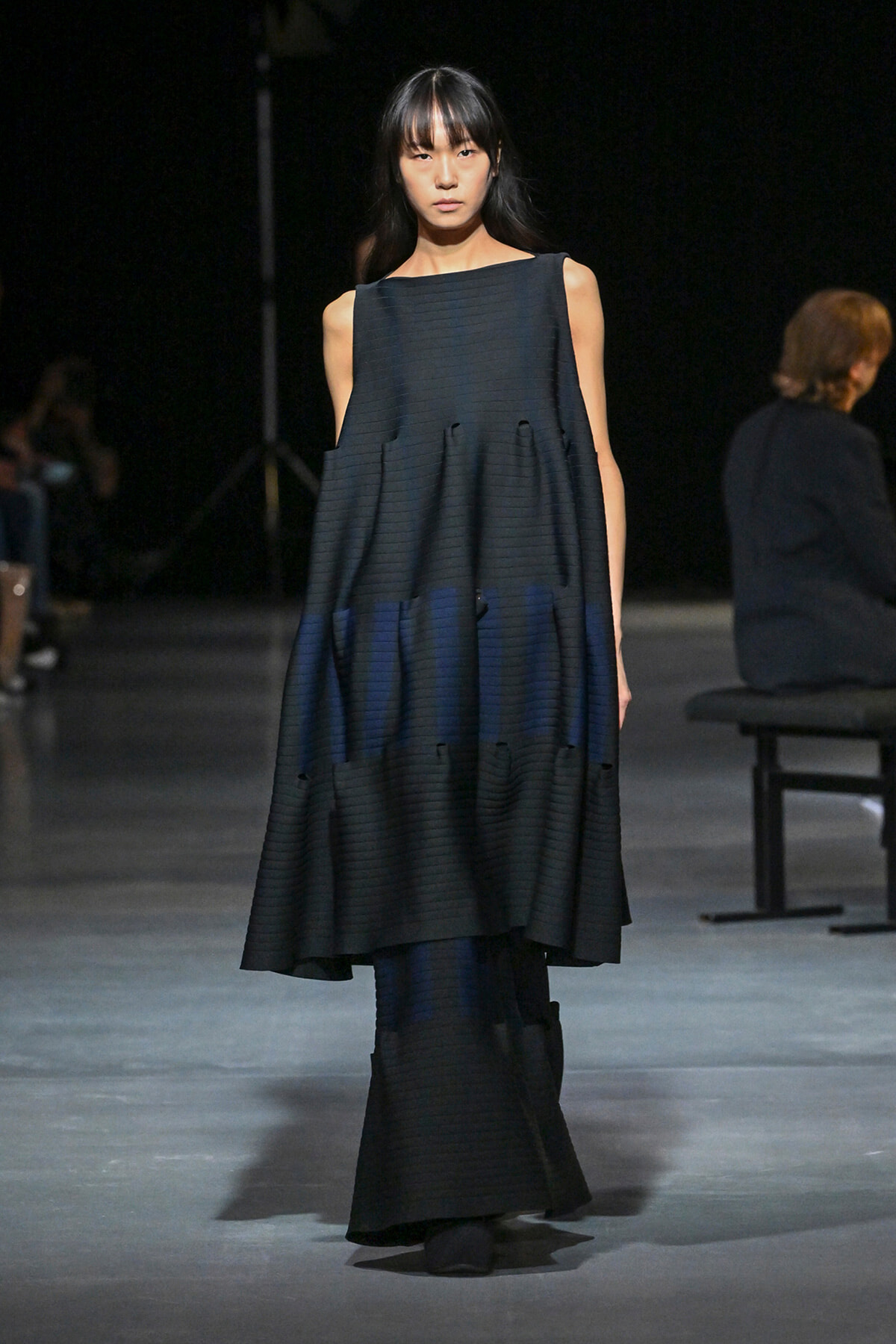 Model on a runway wearing a sleeveless, dark navy tiered maxi dress with subtle horizontal pleats.