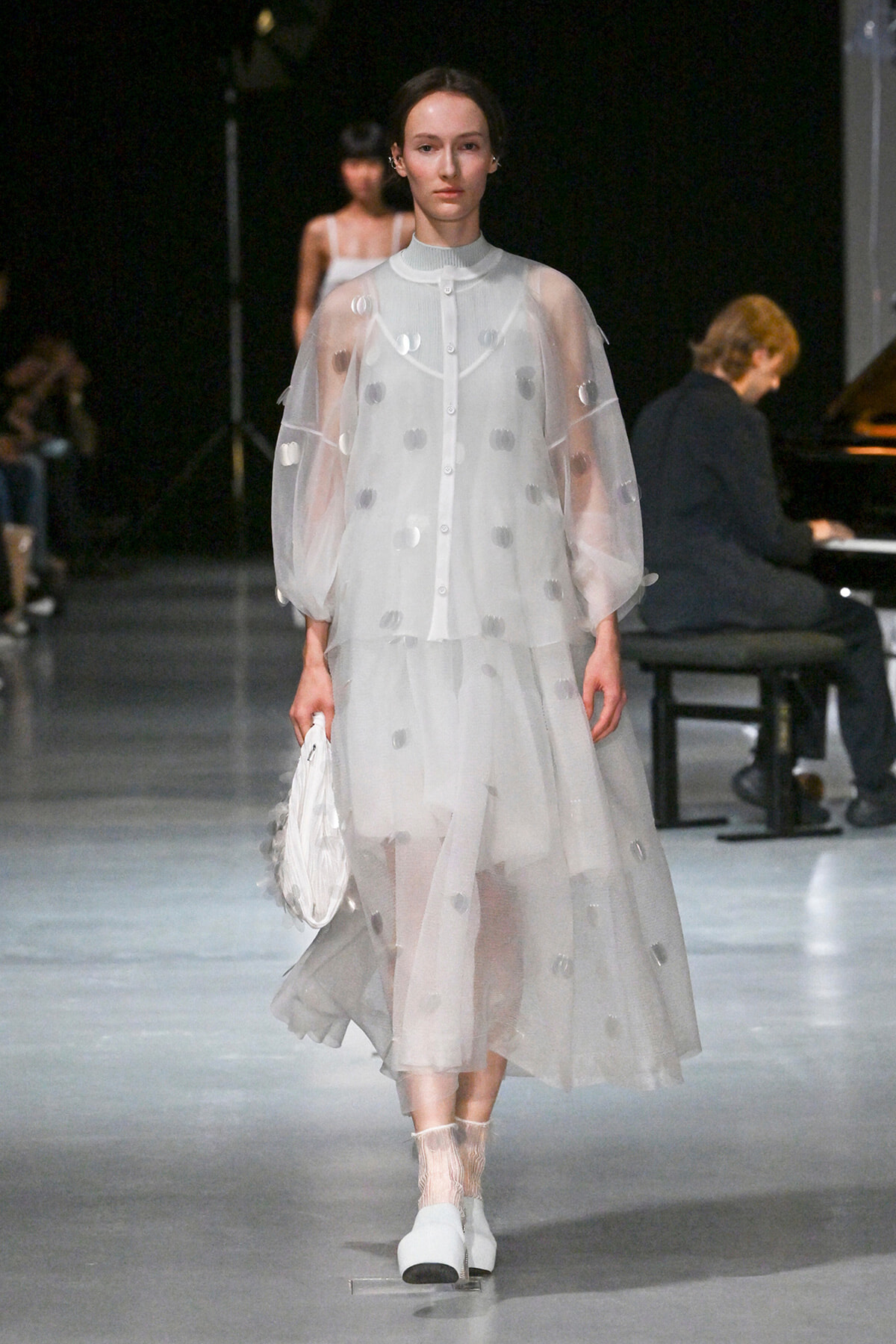 Runway model wears a sheer white polka-dot gown with voluminous sleeves, carrying a white handbag, as a pianist performs in the background.