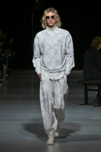 Model on runway wearing pale gray-blue tie-dye oversized top and matching baggy pants, white sneakers, round sunglasses.