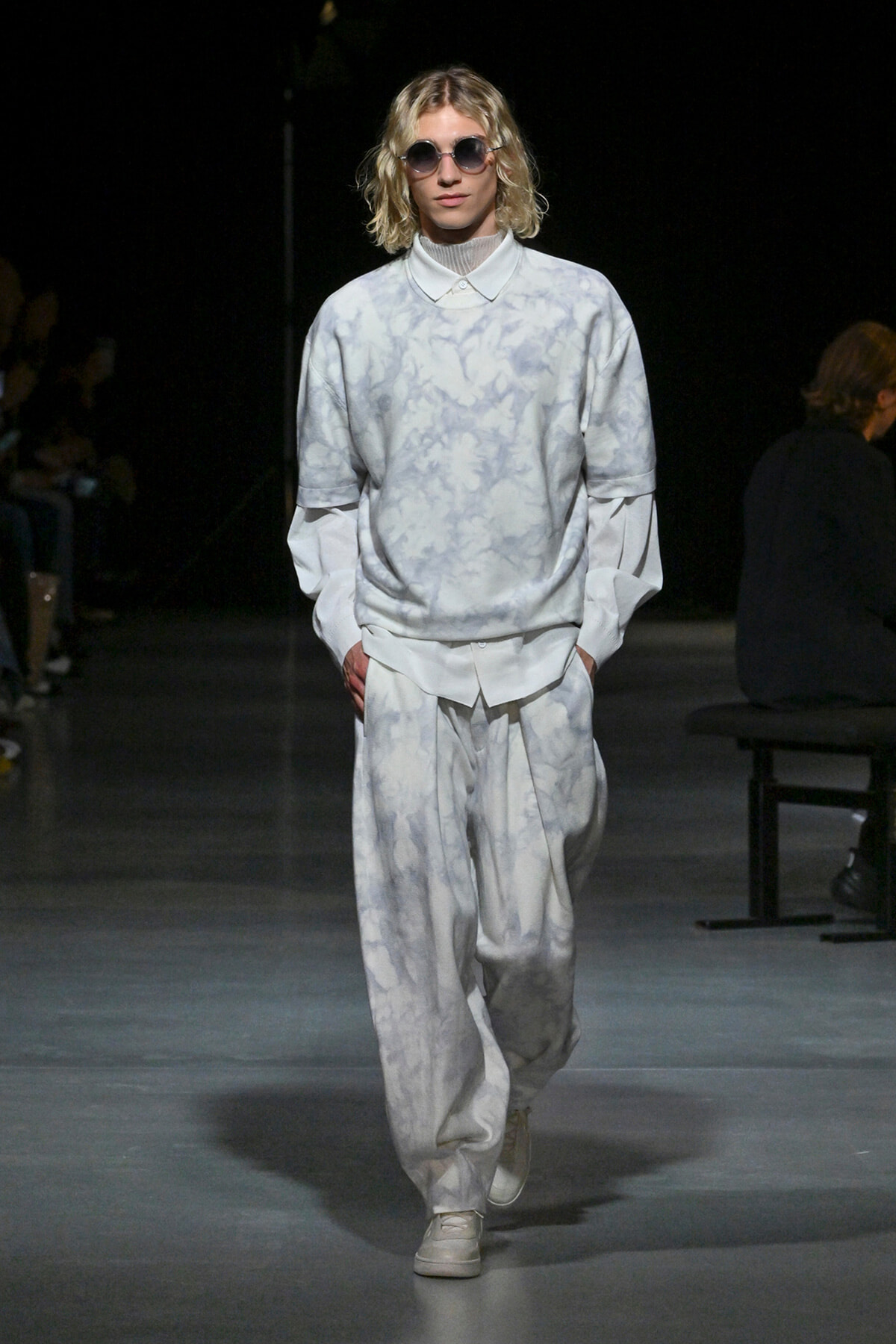 Model on runway wearing pale gray-blue tie-dye oversized top and matching baggy pants, white sneakers, round sunglasses.