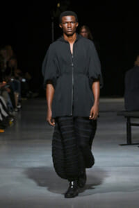 Male model walks a runway in a dark gray zip-front tunic with puff sleeves and a voluminous black textured skirt, black boots beneath.