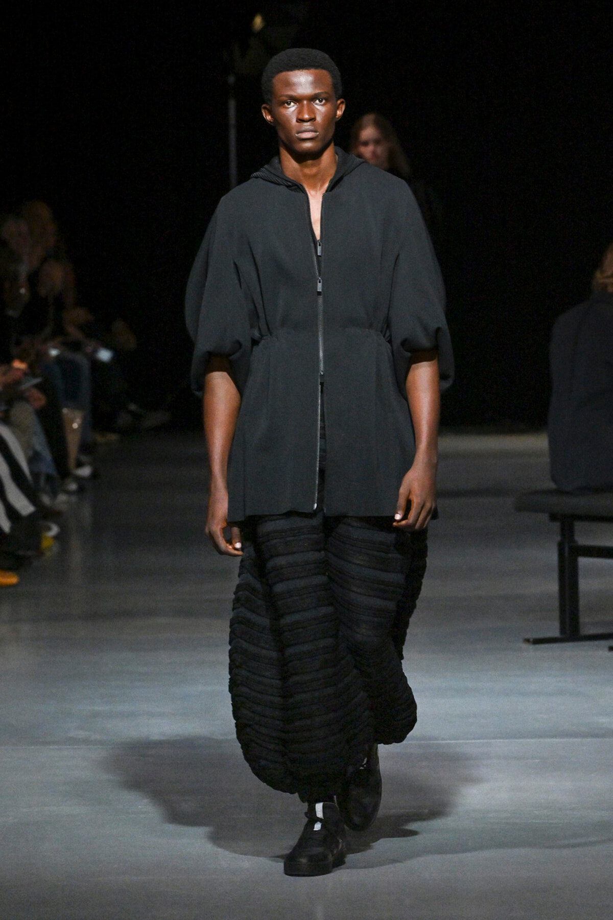 Male model walks a runway in a dark gray zip-front tunic with puff sleeves and a voluminous black textured skirt, black boots beneath.