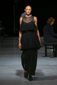 Model walking a runway in a black sleeveless, layered dress with a sheer, striped neckline and loose skirt.