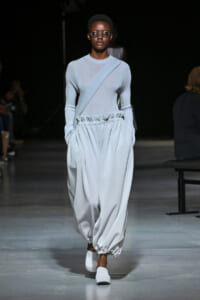 Model walks the runway in a pale blue knit top with a diagonal crossbody sash and oversized gathered trousers, white shoes, and round sunglasses.