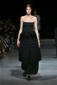 Model walks the runway in a black strapless dress with a large, tiered ruffle skirt; audience on both sides is blurred in the background.
