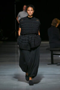 Model walks a runway in an all-black ensemble with a sculpted, horizontally striped textured top and voluminous, cropped black pants.