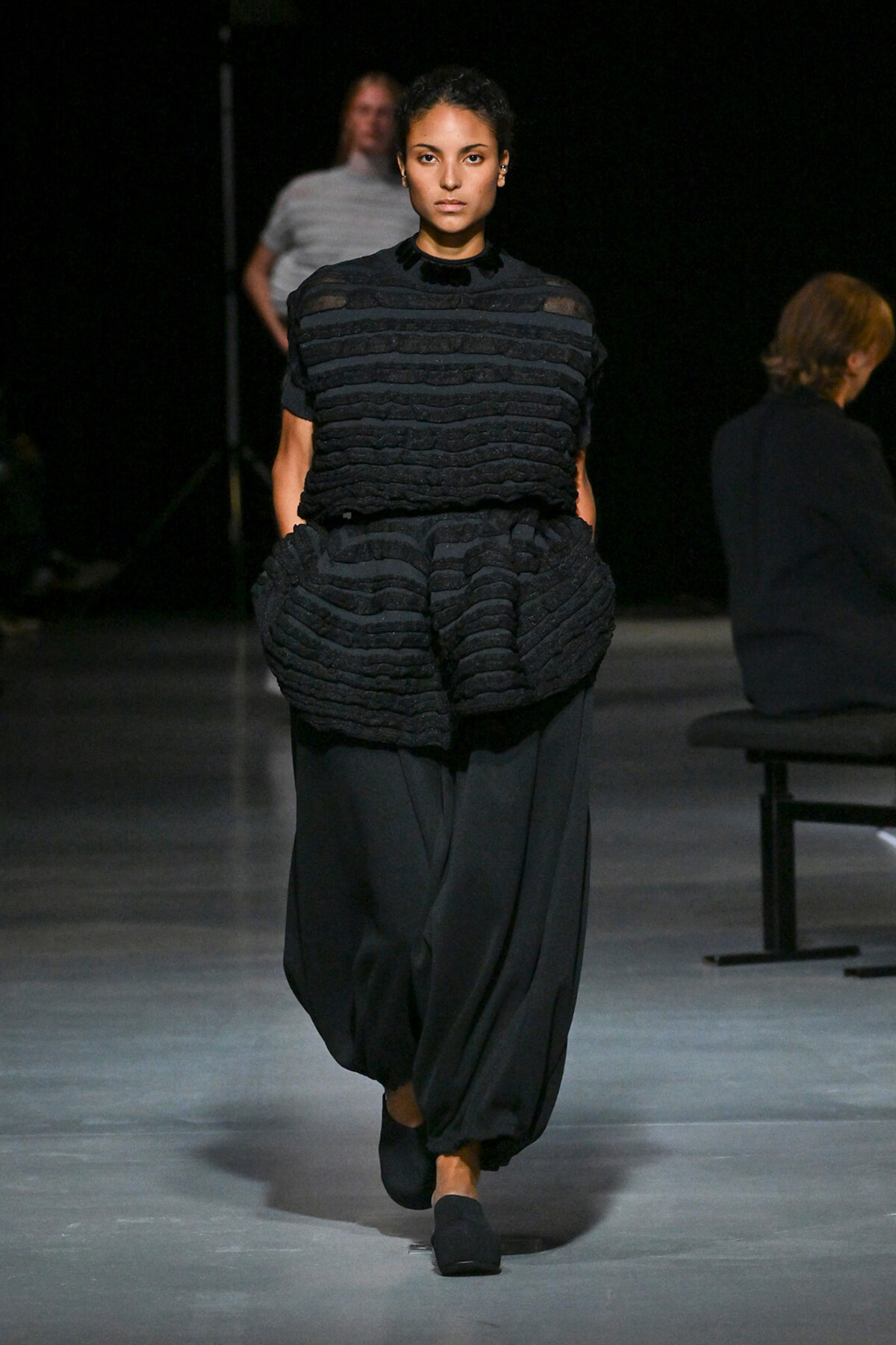 Model walks a runway in an all-black ensemble with a sculpted, horizontally striped textured top and voluminous, cropped black pants.