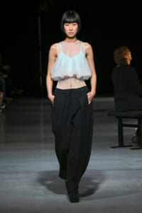 Runway model in a pale blue sheer sleeveless top over a mesh bodice and loose black trousers, walking forward with hands in pockets.