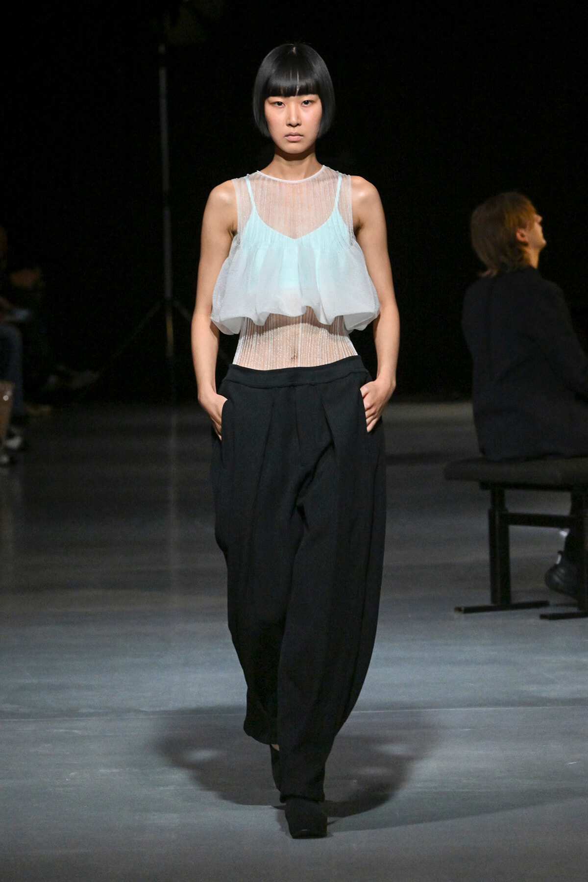 Runway model in a pale blue sheer sleeveless top over a mesh bodice and loose black trousers, walking forward with hands in pockets.