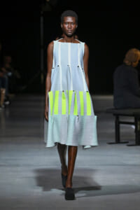 Model walks a fashion runway in a sleeveless light-blue pleated dress with neon yellow panels at the waist.