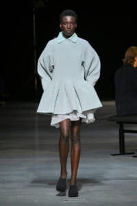 Runway model wearing an oversized pale blue knit sweater-dress with a pale collar, voluminous sleeves, and a layered, pleated hem over a white underdress.