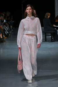Model walking a fashion runway in a sheer pastel pink hoodie and matching wide-leg pants, carrying a pink bag.