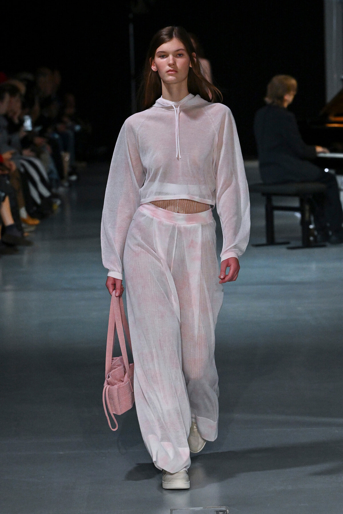 Model walking a fashion runway in a sheer pastel pink hoodie and matching wide-leg pants, carrying a pink bag.