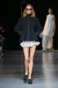 Model on a fashion runway wearing an oversized black zip-front jacket, a white layered mini skirt, black round sunglasses, and black slip-on shoes.