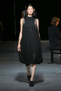 Model walks the runway in a black, sleeveless midi dress with sheer panels and a high neckline.