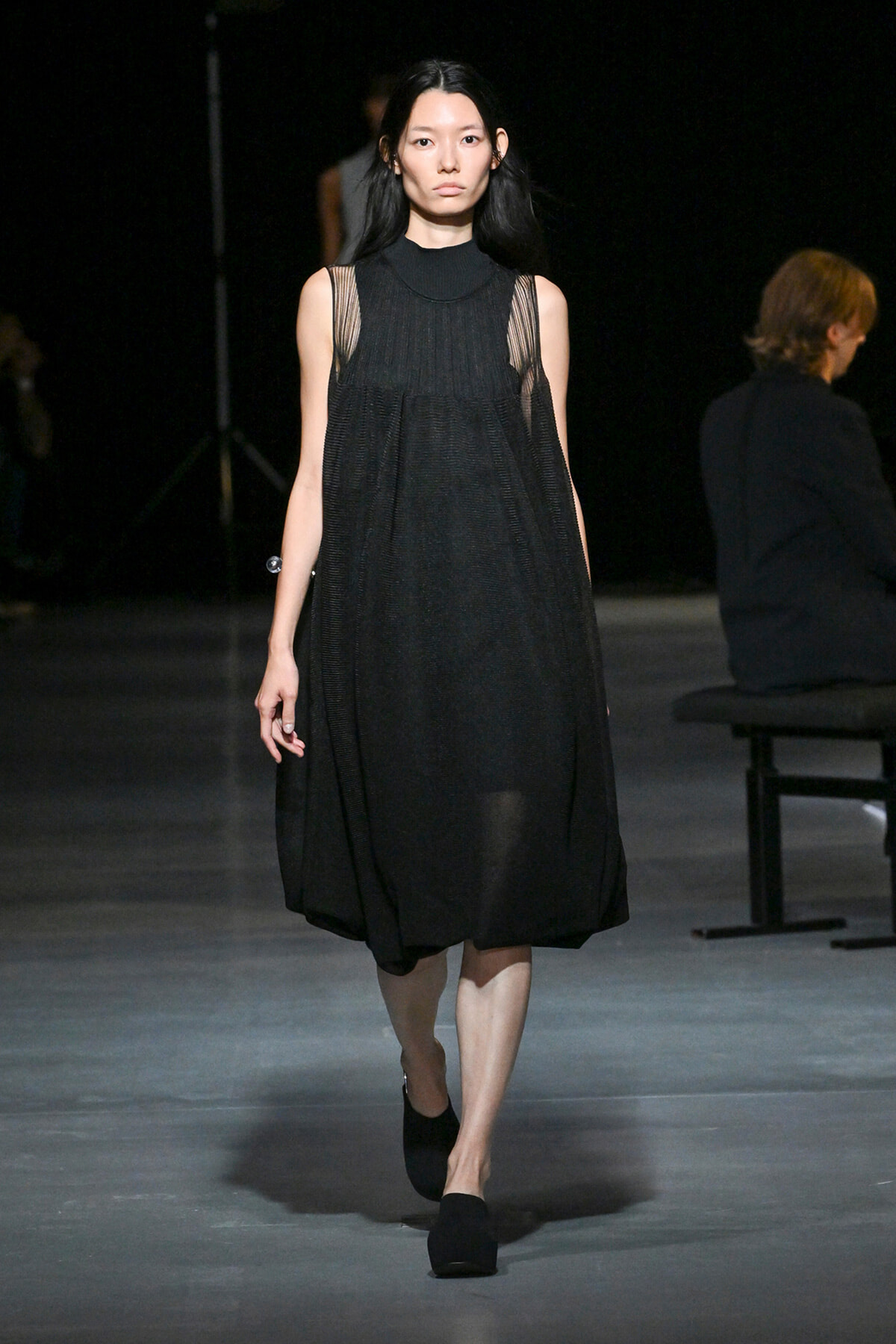Model walks the runway in a black, sleeveless midi dress with sheer panels and a high neckline.