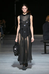 Model on a runway wearing a sheer black sleeveless maxi dress with circular disc embellishments, holding a matching black bag.