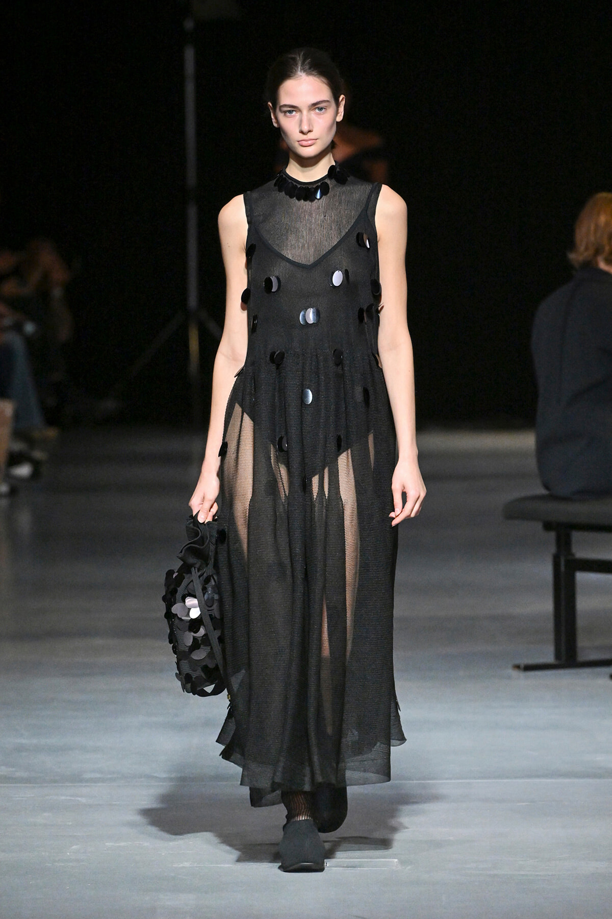 Model on a runway wearing a sheer black sleeveless maxi dress with circular disc embellishments, holding a matching black bag.