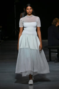 Model on a runway wearing a pale blue, knee/tea-length dress with a fitted bodice and a layered, sheer-skirt bottom and short sleeves.