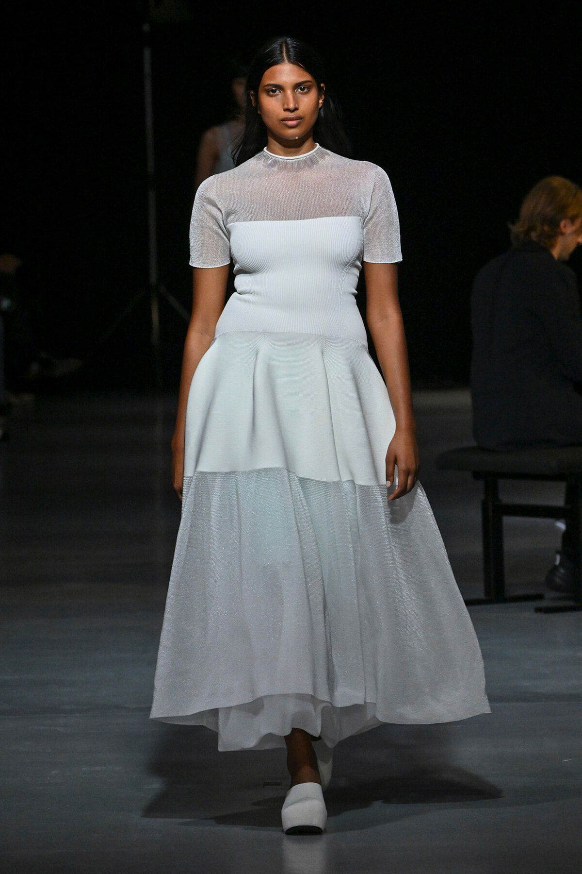 Model on a runway wearing a pale blue, knee/tea-length dress with a fitted bodice and a layered, sheer-skirt bottom and short sleeves.