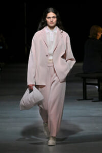 Model on a runway wearing a loose pale pink suit with a white polo shirt, carrying a white striped bag.