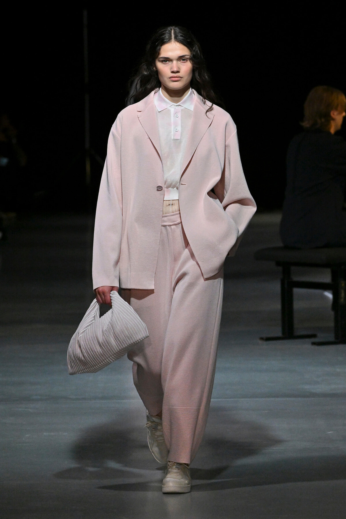 Model on a runway wearing a loose pale pink suit with a white polo shirt, carrying a white striped bag.