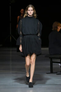 Model walking a fashion runway in a black, high-neck dress with sheer puff sleeves.