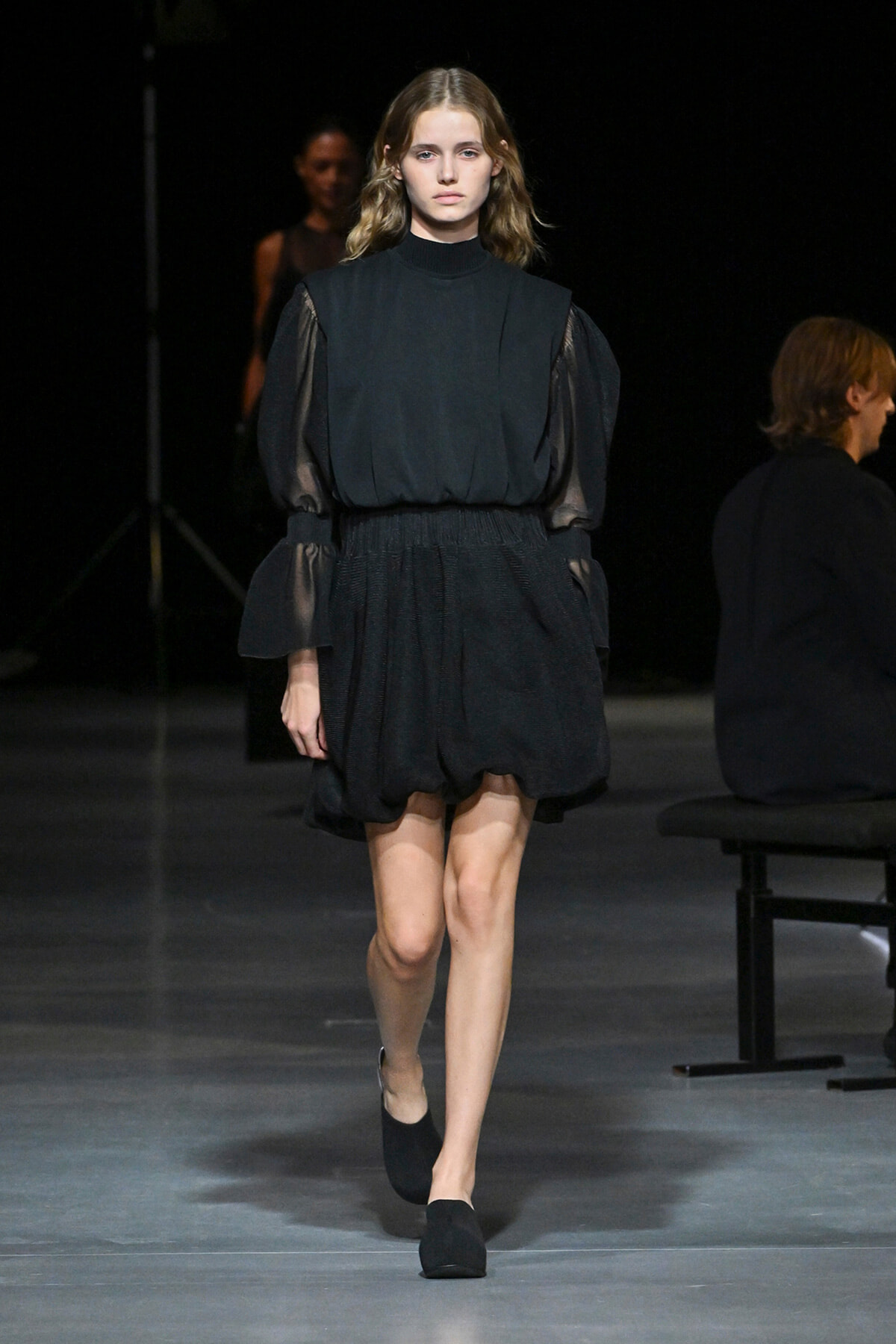Model walking a fashion runway in a black, high-neck dress with sheer puff sleeves.