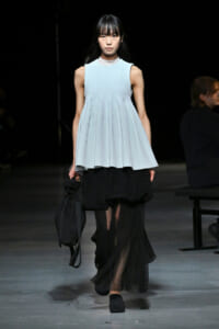 Model walking runway in a light blue sleeveless peplum top over a black sheer skirt, carrying a black bag.