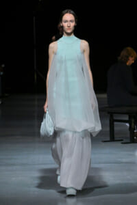 Runway model walking in a pale blue, layered sheer dress with a high neckline, holding a light-blue striped clutch.