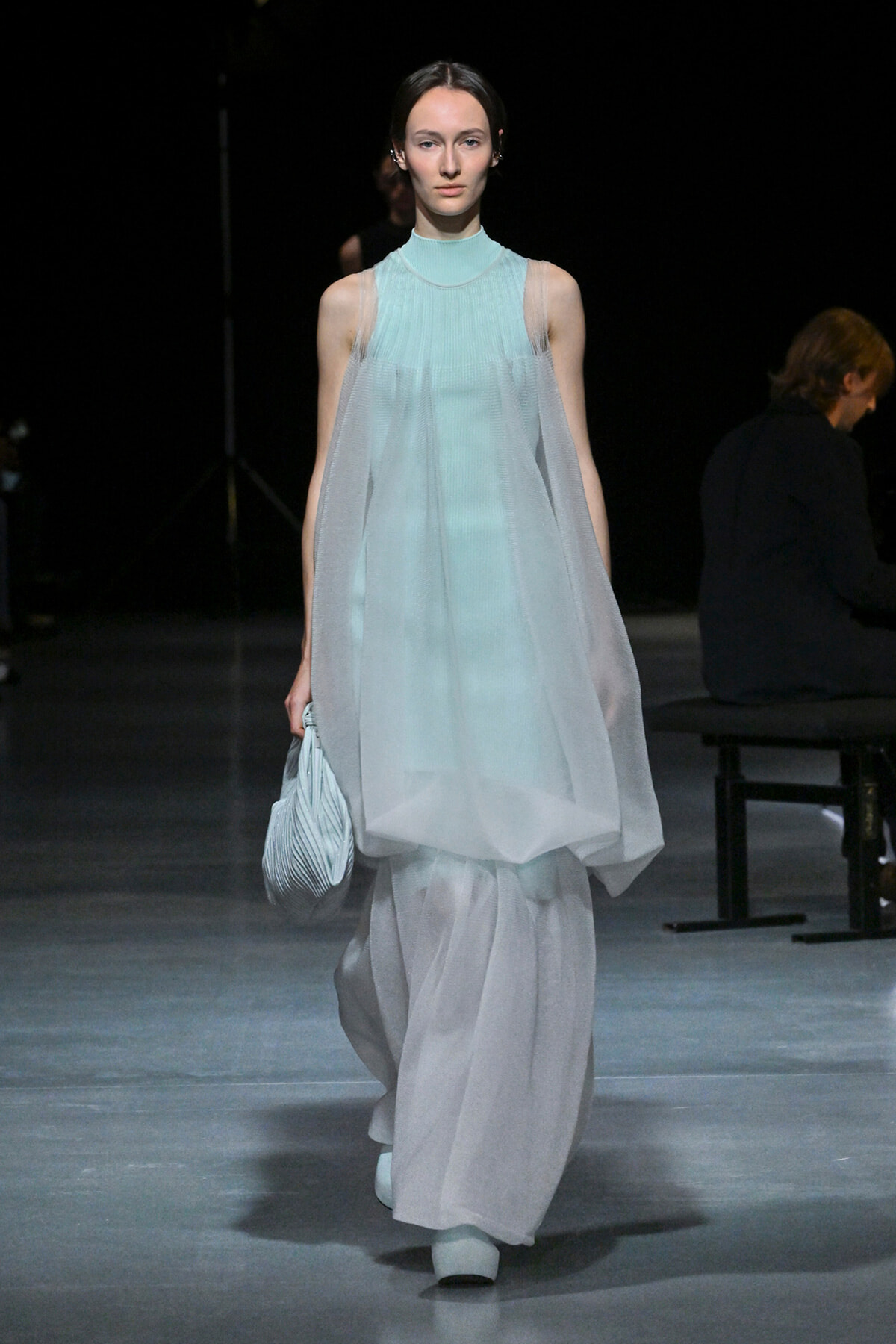 Runway model walking in a pale blue, layered sheer dress with a high neckline, holding a light-blue striped clutch.