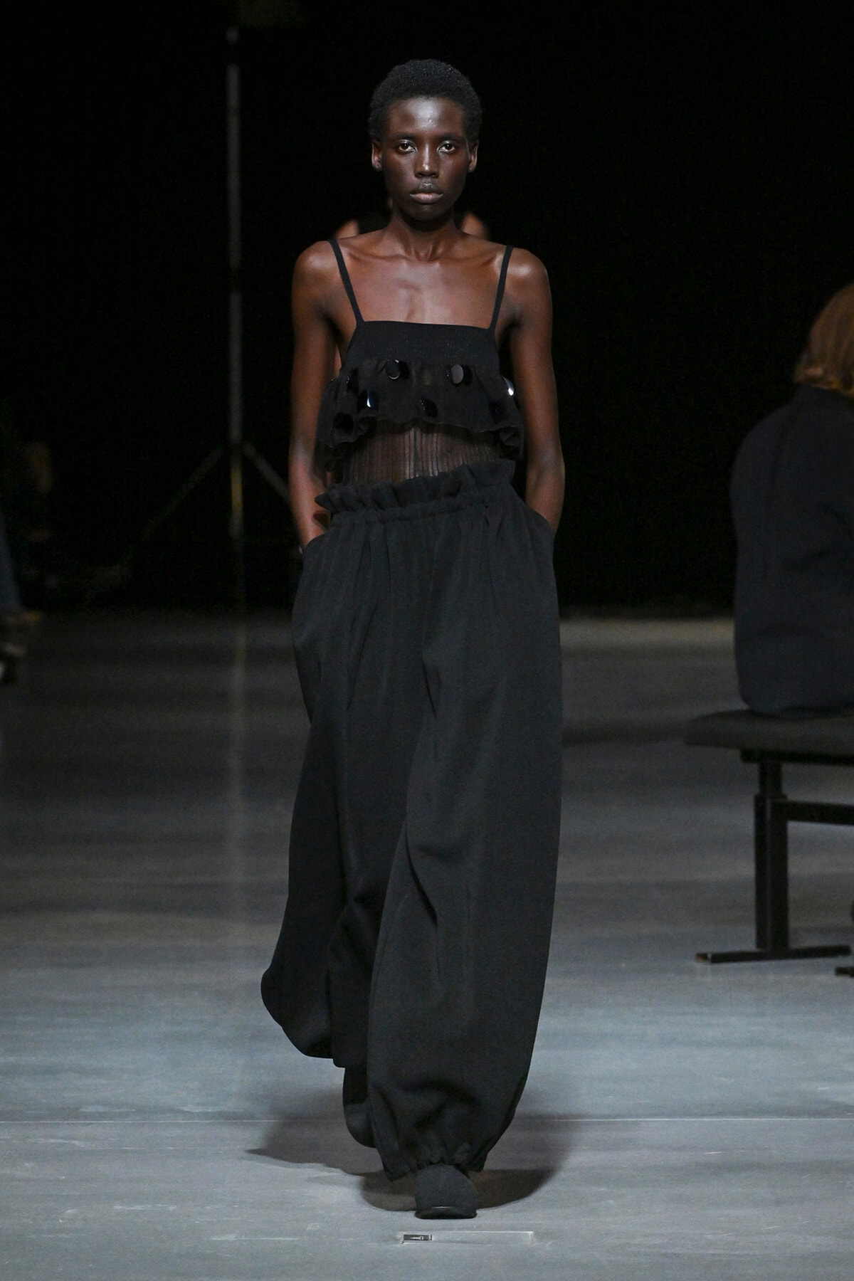 Runway model walks in an all-black outfit: sheer, ruffled top with thin straps and wide, billowy pants.