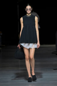 Model on a runway wearing a sleeveless dark pleated tunic over a white ruffled skirt, round glasses, and black pleated handbag.