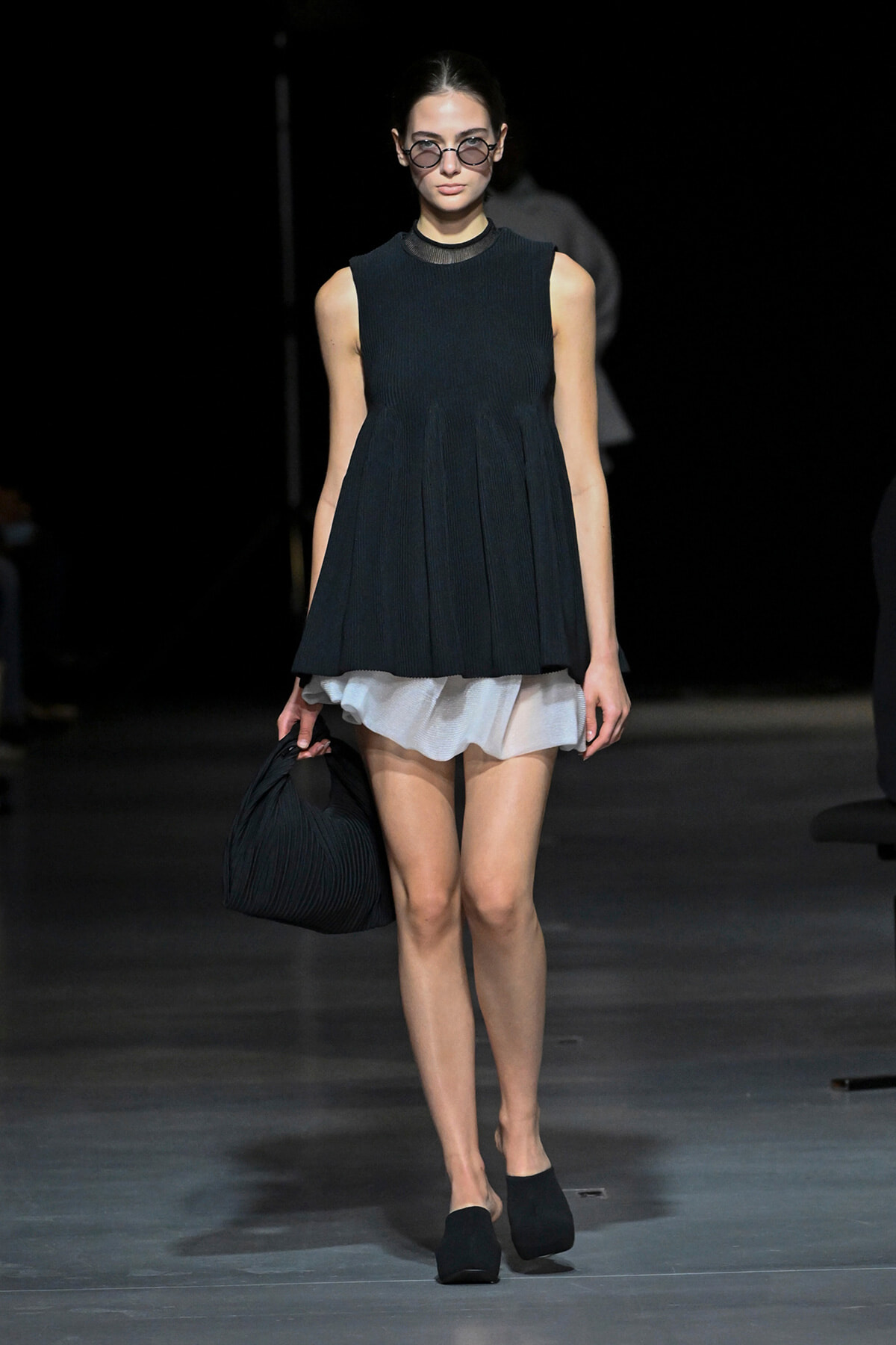 Model on a runway wearing a sleeveless dark pleated tunic over a white ruffled skirt, round glasses, and black pleated handbag.