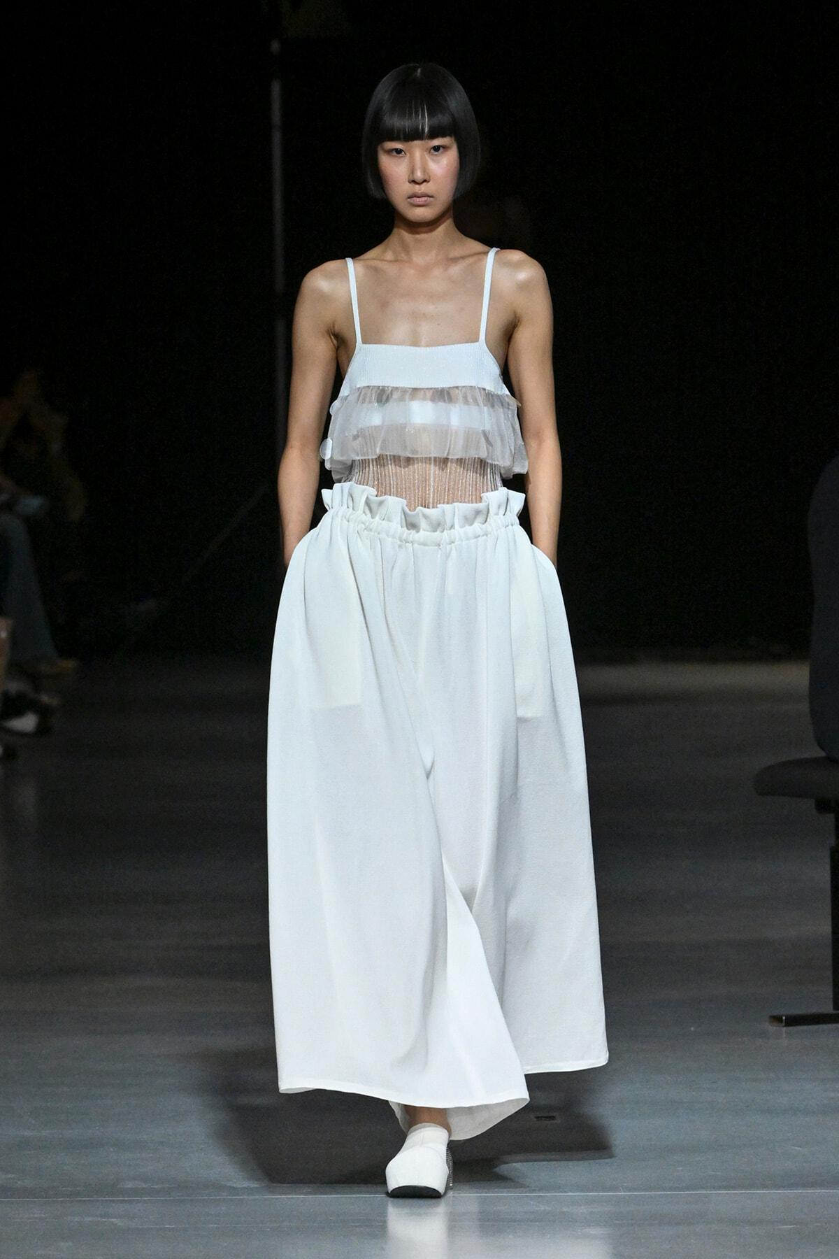 Model walking a runway in a pale blue sheer-strap top with layered tulle and a high-waisted flowing skirt, white shoes.