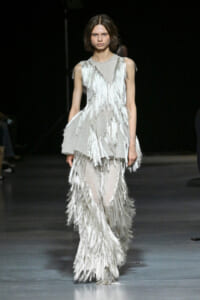 Model walks the runway in a silver fringe gown with cascading tassels on a dark catwalk